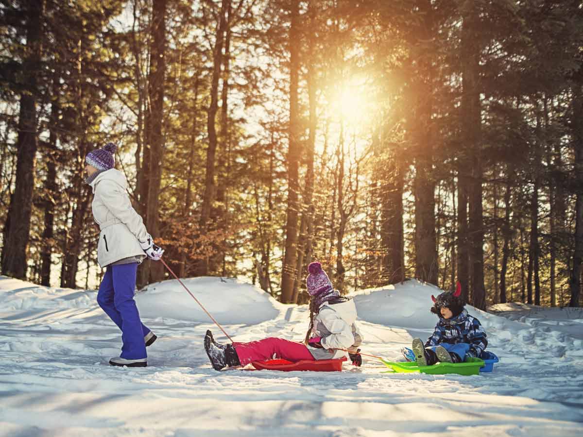 Jeune mère tirant 2 traineaux où prennent place 2 enfants lors d’une promenade hivernale en forêt.